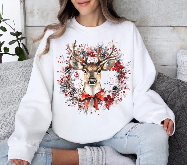 May include: White sweatshirt featuring a watercolor illustration of a deer with antlers, a red bow, and a wreath of pine needles, pine cones, and red flowers. The design is centered on the front of the sweatshirt.