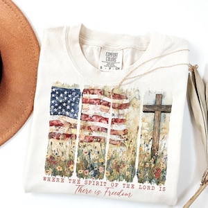 May include: A beige t-shirt with a watercolor design of three American flags and a wooden cross. The flags are surrounded by wildflowers. The text "Where the Spirit of the Lord is There is Freedom" is printed below the design.