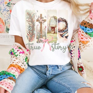 Puede incluir: Camiseta blanca con un dise&ntilde;o gr&aacute;fico de una cruz, una corona de espinas, una piedra, una hoja de palma y las palabras "true story".