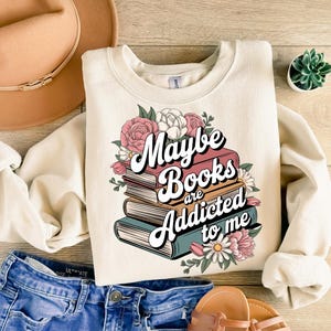 May include: A cream-colored sweatshirt with a graphic of a stack of books with flowers and the text "Maybe Books are Addicted to me".