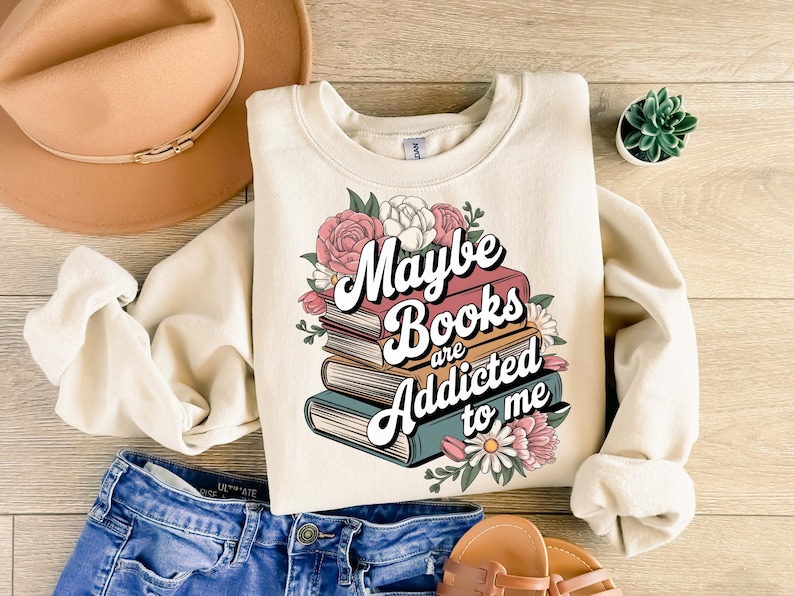 May include: A cream-colored sweatshirt with a graphic of a stack of books with flowers and the text "Maybe Books are Addicted to me".