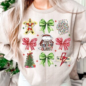 May include: Beige sweatshirt with a festive Christmas-themed design. The design includes images of a Christmas tree, bows, Santa Claus, and a camper with a lightning bolt. Text on the shirt reads "You Serious Clark!"