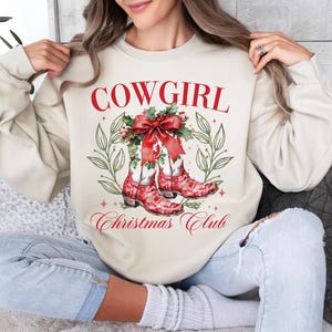 Op de afbeelding: Een cr&egrave;mekleurige sweatshirt met de tekst "COWGIRL Christmas Club" in rode letters. Het ontwerp toont rode cowboylaarzen met een kerstthema, versierd met een rode strik en hulst. De sweatshirt is een casual, feestelijk kledingstuk.