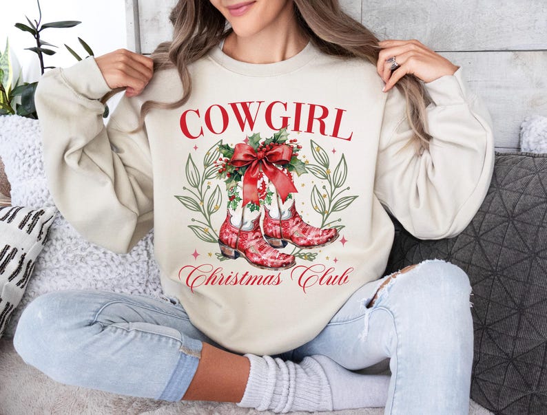 Op de afbeelding: Een cr&egrave;mekleurige sweatshirt met de tekst "COWGIRL Christmas Club" in rode letters. Het ontwerp toont rode cowboylaarzen met een kerstthema, versierd met een rode strik en hulst. De sweatshirt is een casual, feestelijk kledingstuk.