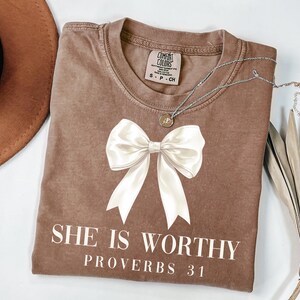 May include: A brown t-shirt with a large white bow graphic and the text "SHE IS WORTHY PROVERBS 31". The shirt has a crew neck and short sleeves. A silver necklace with a small pendant is on the shirt.