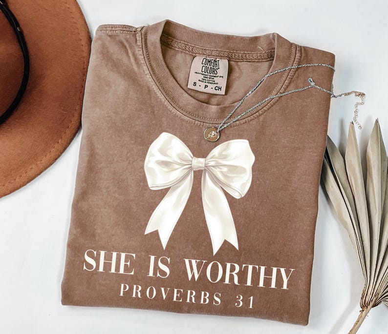 May include: A brown t-shirt with a large white bow graphic and the text "SHE IS WORTHY PROVERBS 31". The shirt has a crew neck and short sleeves. A silver necklace with a small pendant is on the shirt.