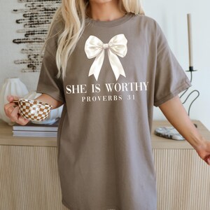 May include: A brown t-shirt with a large white bow graphic and the text "SHE IS WORTHY PROVERBS 31" in a serif font. The shirt has short sleeves and a relaxed fit.