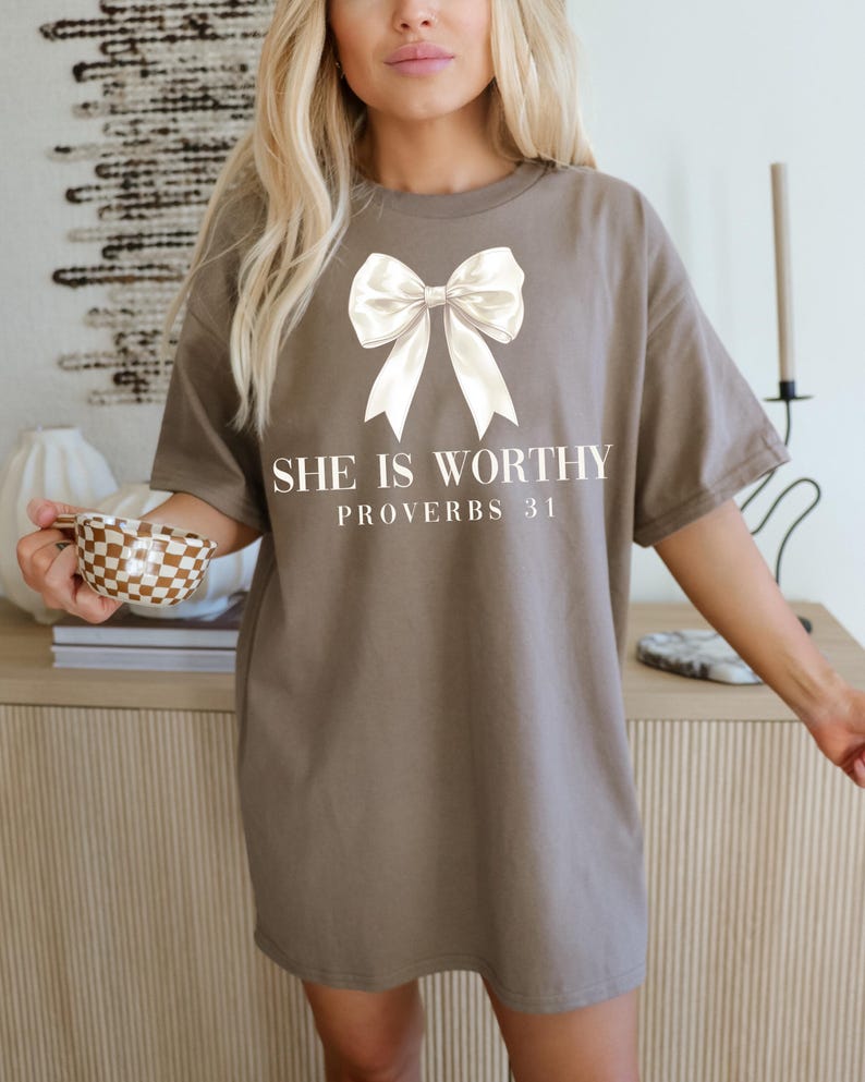 May include: A brown t-shirt with a large white bow graphic and the text "SHE IS WORTHY PROVERBS 31" in a serif font. The shirt has short sleeves and a relaxed fit.