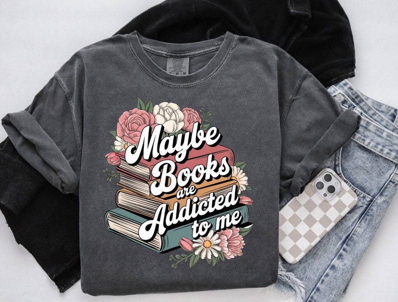 May include: A dark gray t-shirt with a stack of books and flowers. The text on the shirt reads "Maybe books are addicted to me".