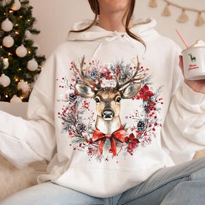 May include: White hoodie featuring a watercolor illustration of a deer with large antlers, surrounded by a wreath of red flowers, pine cones, and greenery. A red bow is at the bottom. The hoodie is a festive Christmas design.