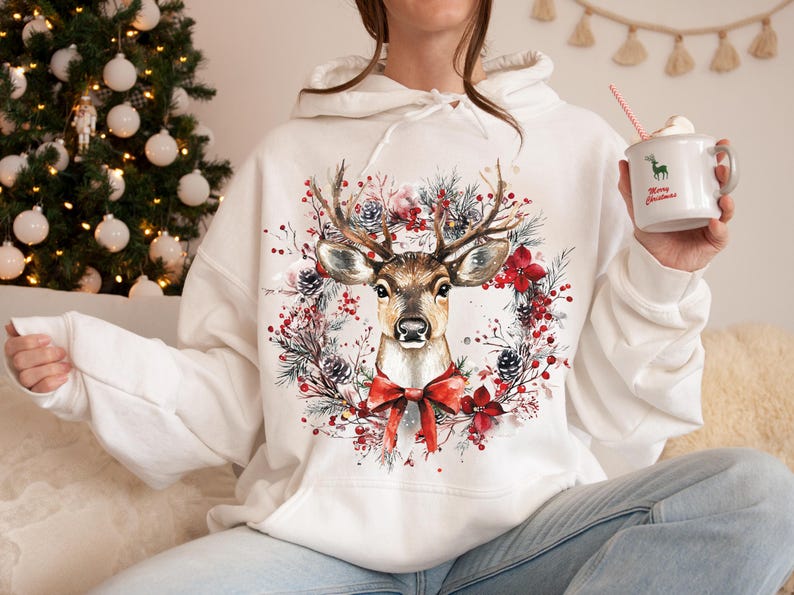 May include: White hoodie featuring a watercolor illustration of a deer with large antlers, surrounded by a wreath of red flowers, pine cones, and greenery. A red bow is at the bottom. The hoodie is a festive Christmas design.