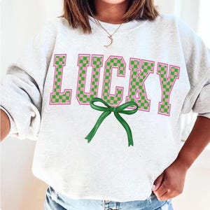 May include: A white sweatshirt with the word "LUCKY" printed in large, pink and green checkered letters. A green bow is centered below the word.