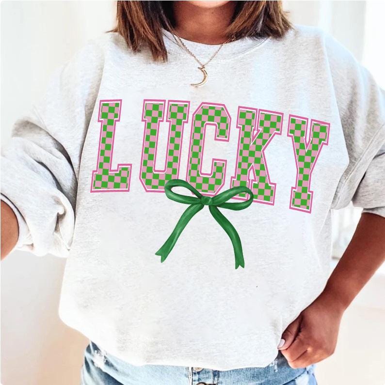 May include: A white sweatshirt with the word "LUCKY" printed in large, pink and green checkered letters. A green bow is centered below the word.