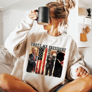 May include: A white sweatshirt with the text "That's My President" and images of three men in front of an American flag.