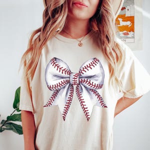 Pu&ograve; includere: T-shirt color crema con un design a fiocco ad acquerello. Il fiocco &egrave; progettato per assomigliare a una palla da baseball, con sezioni bianche e cuciture rosse. La maglietta &egrave; uno stile casual e confortevole.