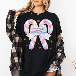 May include: Black t-shirt featuring a large, iridescent candy cane design with a bow and gold accents. The candy canes have a plaid pattern in pastel colors. Gold snowflake accents are scattered around the design.