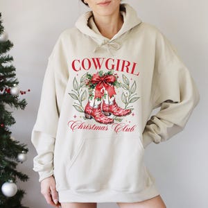 Op de afbeelding: Een beige hoodie met een grafische print op de voorkant. De print toont rode tekst "COWGIRL" boven een aquarel illustratie van rode cowboylaarzen versierd met een strik en groen. Onder de laarzen staat de tekst "Christmas Club".