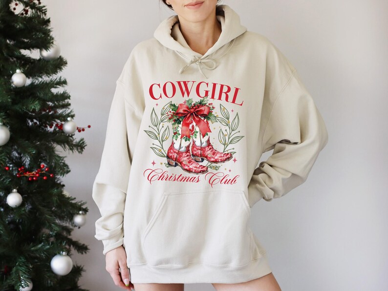 Op de afbeelding: Een beige hoodie met een grafische print op de voorkant. De print toont rode tekst "COWGIRL" boven een aquarel illustratie van rode cowboylaarzen versierd met een strik en groen. Onder de laarzen staat de tekst "Christmas Club".