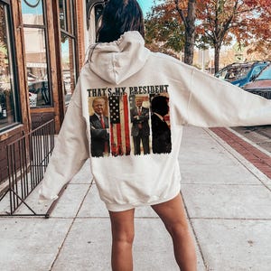 May include: White hooded sweatshirt with a graphic print of three presidents in front of an American flag. The text "That's My President" is printed above the graphic.