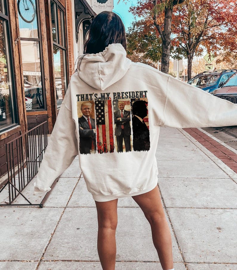 May include: White hooded sweatshirt with a graphic print of three presidents in front of an American flag. The text "That's My President" is printed above the graphic.