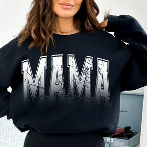 Op de afbeelding: Zwart sweatshirt met een witte, verweerde grafische afbeelding met de tekst "MAMA" in grote blokletters.