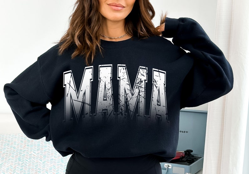 Op de afbeelding: Zwart sweatshirt met een witte, verweerde grafische afbeelding met de tekst "MAMA" in grote blokletters.