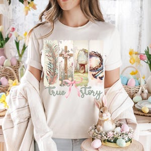 Puede incluir: Una camiseta blanca con un dise&ntilde;o gr&aacute;fico de una cruz, una corona de espinas, una tumba y una hoja de palma. El texto "true story" est&aacute; impreso debajo del gr&aacute;fico.
