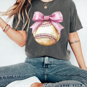 May include: A charcoal grey t-shirt with a pink bow on a baseball. The baseball is yellow with red stitching.