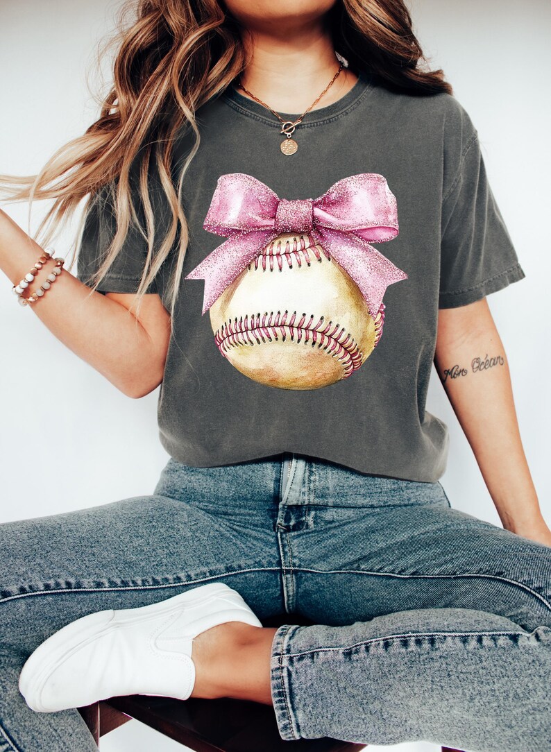 May include: A charcoal grey t-shirt with a pink bow on a baseball. The baseball is yellow with red stitching.