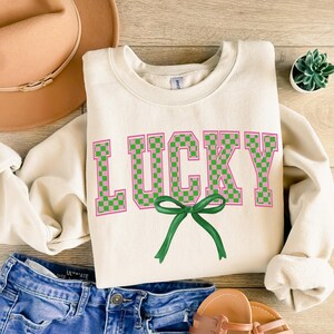 May include: A cream-colored sweatshirt with the word "LUCKY" in pink and green checkered letters. A green bow is centered below the word.