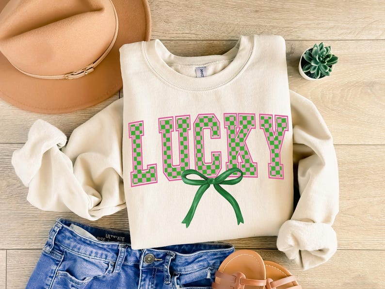 May include: A cream-colored sweatshirt with the word "LUCKY" in pink and green checkered letters. A green bow is centered below the word.