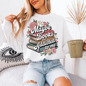 May include: White sweatshirt with a graphic print of stacked books and flowers. The text reads "Maybe books are addicted to me".