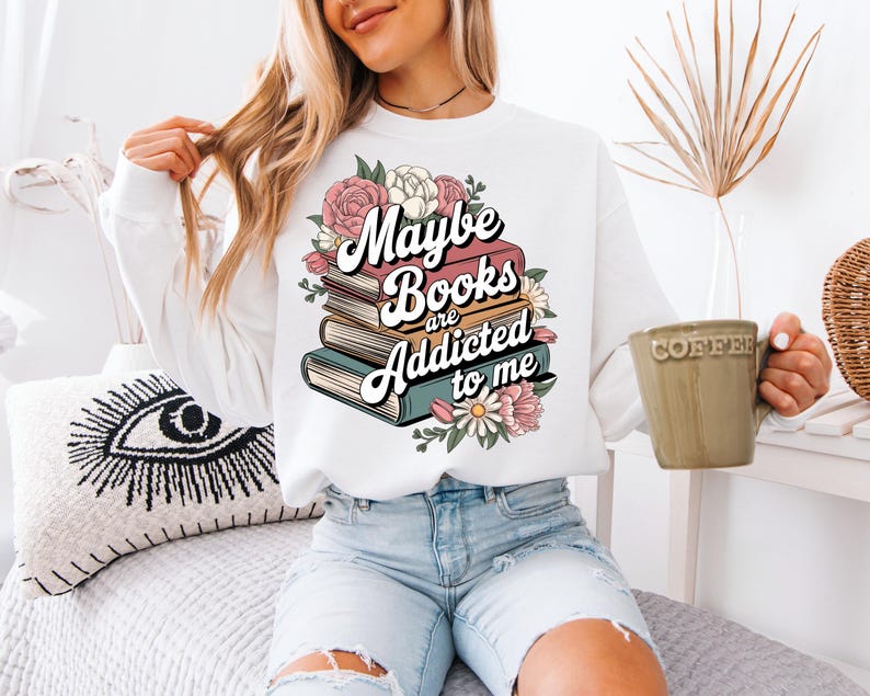 May include: White sweatshirt with a graphic print of stacked books and flowers. The text reads "Maybe books are addicted to me".