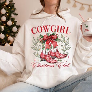 Op de afbeelding: Witte hoodie met een rode "COWGIRL" afbeelding, met rode cowboylaarzen, een strik en de tekst "Christmas Club". Het ontwerp bevat groene bladeren en sterren. Een mok met een hert en "Merry Christmas" wordt vastgehouden.