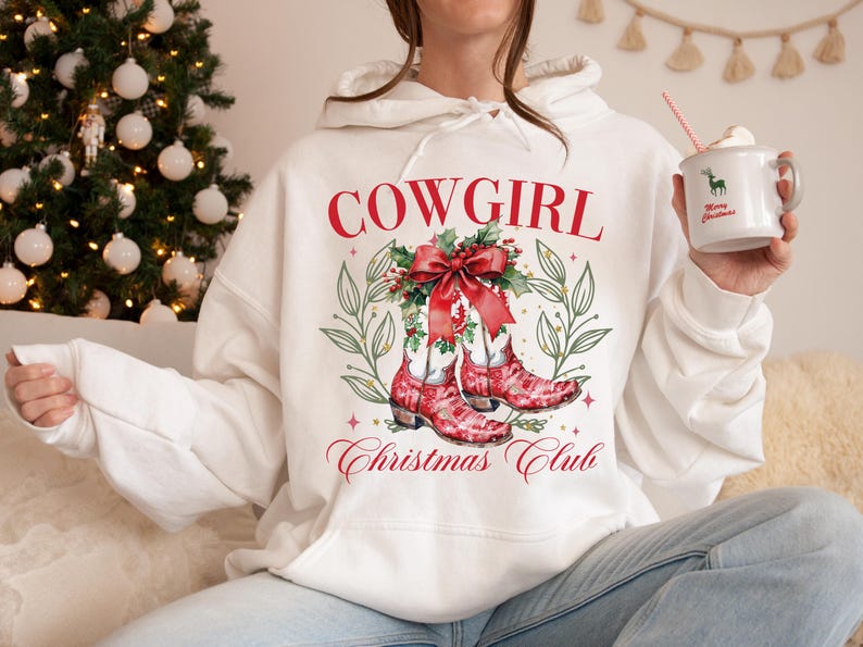 Op de afbeelding: Witte hoodie met een rode "COWGIRL" afbeelding, met rode cowboylaarzen, een strik en de tekst "Christmas Club". Het ontwerp bevat groene bladeren en sterren. Een mok met een hert en "Merry Christmas" wordt vastgehouden.