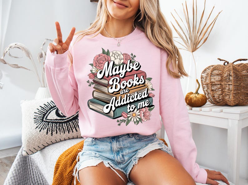 May include: A pink sweatshirt with a graphic design of a stack of books with flowers and the text "Maybe books are addicted to me".
