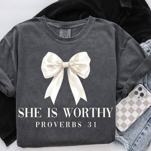 May include: Dark gray t-shirt with a large white bow graphic. The text "SHE IS WORTHY PROVERBS 31" is printed below the bow. The shirt is paired with blue jeans and a phone case with a checkered pattern.