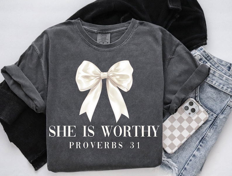 May include: Dark gray t-shirt with a large white bow graphic. The text "SHE IS WORTHY PROVERBS 31" is printed below the bow. The shirt is paired with blue jeans and a phone case with a checkered pattern.