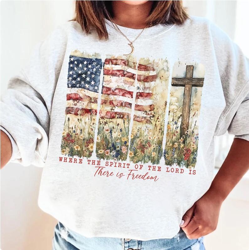May include: A gray sweatshirt with a watercolor design of an American flag, flowers, and a wooden cross. The text "Where the Spirit of the Lord is There is Freedom" is printed below the design.