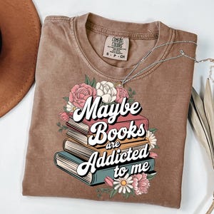 May include: A brown t-shirt with a graphic design of a stack of books with flowers and the text "Maybe Books are Addicted to me".
