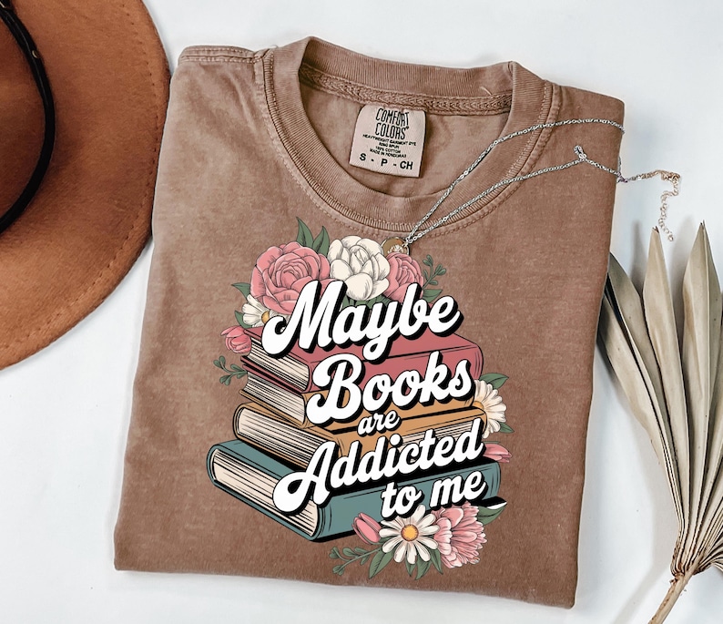 May include: A brown t-shirt with a graphic design of a stack of books with flowers and the text "Maybe Books are Addicted to me".