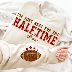 May include: A beige sweatshirt with red glitter text that reads "I'm just here for the halftime show". A brown glitter football with five gold stars is on the sleeve.