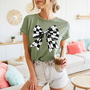 May include: A green t-shirt featuring a black and white checkered bow design. The bow is centrally positioned on the shirt and is the main design element.