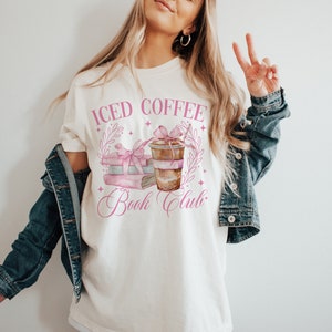 K&ouml;nnte beinhalten: Wei&szlig;es T-Shirt mit einem rosafarbenen Grafikdesign, auf dem "Iced Coffee Book Club" steht. Die Grafik zeigt einen Stapel B&uuml;cher mit einem rosafarbenen Band und einer Tasse Eiskaffee.