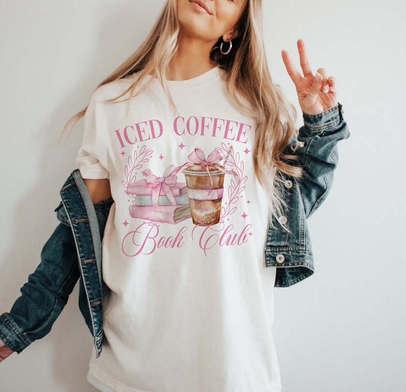 K&ouml;nnte beinhalten: Wei&szlig;es T-Shirt mit einem rosafarbenen Grafikdesign, auf dem "Iced Coffee Book Club" steht. Die Grafik zeigt einen Stapel B&uuml;cher mit einem rosafarbenen Band und einer Tasse Eiskaffee.