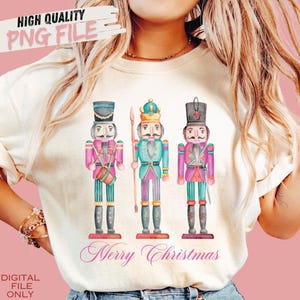 Puede incluir: Una camiseta blanca con un diseño de acuarela de tres cascanueces. Los cascanueces llevan uniformes coloridos y sostienen varios accesorios. El texto "Feliz Navidad" está escrito en rosa debajo de los cascanueces.