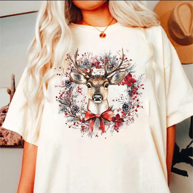 May include: Cream-colored t-shirt featuring a watercolor illustration of a deer's head within a Christmas wreath. The wreath includes pine needles, red berries, and a red bow. The deer has large antlers and a detailed face.