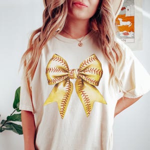 Pu&ograve; includere: T-shirt color crema con un grande fiocco in stile acquerello. Il fiocco &egrave; giallo con cuciture marroni, che ricordano una palla da baseball. Il design &egrave; centrato sulla maglietta, creando un look sportivo ed elegante.