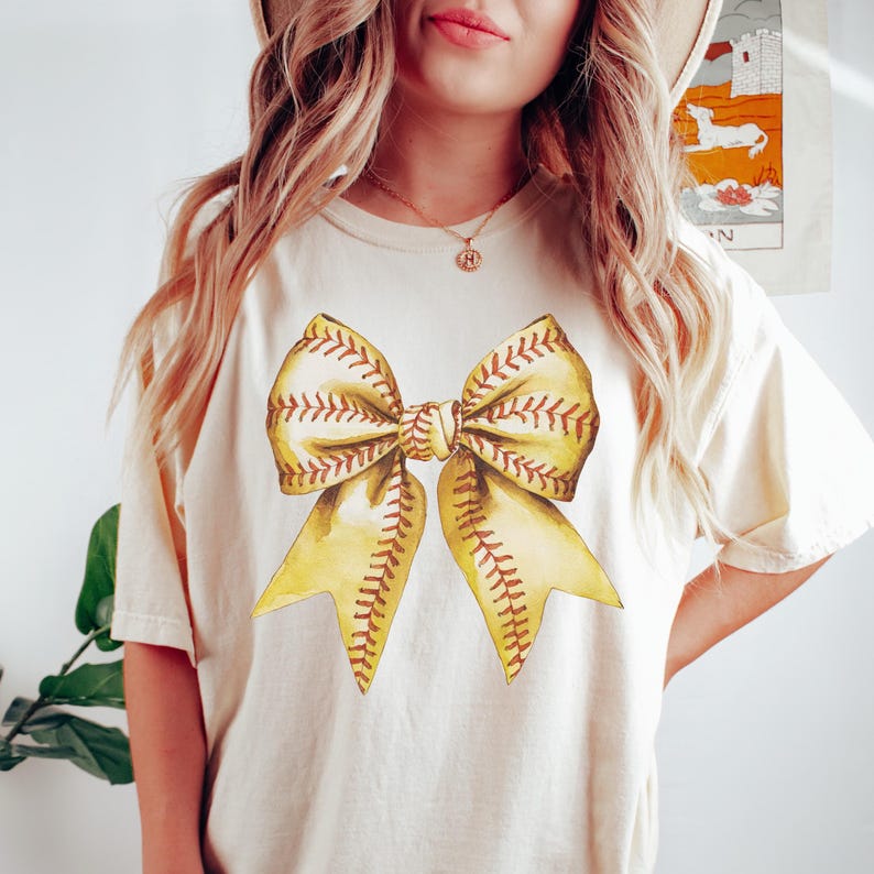 Pu&ograve; includere: T-shirt color crema con un grande fiocco in stile acquerello. Il fiocco &egrave; giallo con cuciture marroni, che ricordano una palla da baseball. Il design &egrave; centrato sulla maglietta, creando un look sportivo ed elegante.