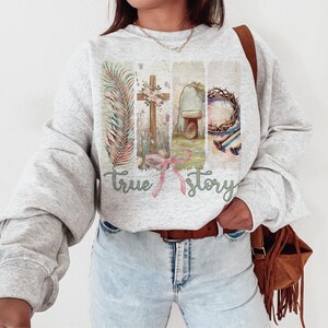 Puede incluir: Una sudadera gris con un estampado gr&aacute;fico de una cruz, una corona de espinas y una tumba. El texto "true story" est&aacute; impreso debajo del gr&aacute;fico.